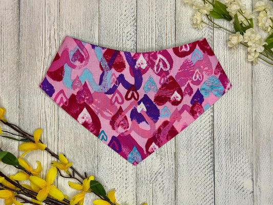 A pet bandana with a pink background featuring varying colors of hearts (pink, red, white, purple, blue) displayed on a wooden surface with flowers for decoration. 