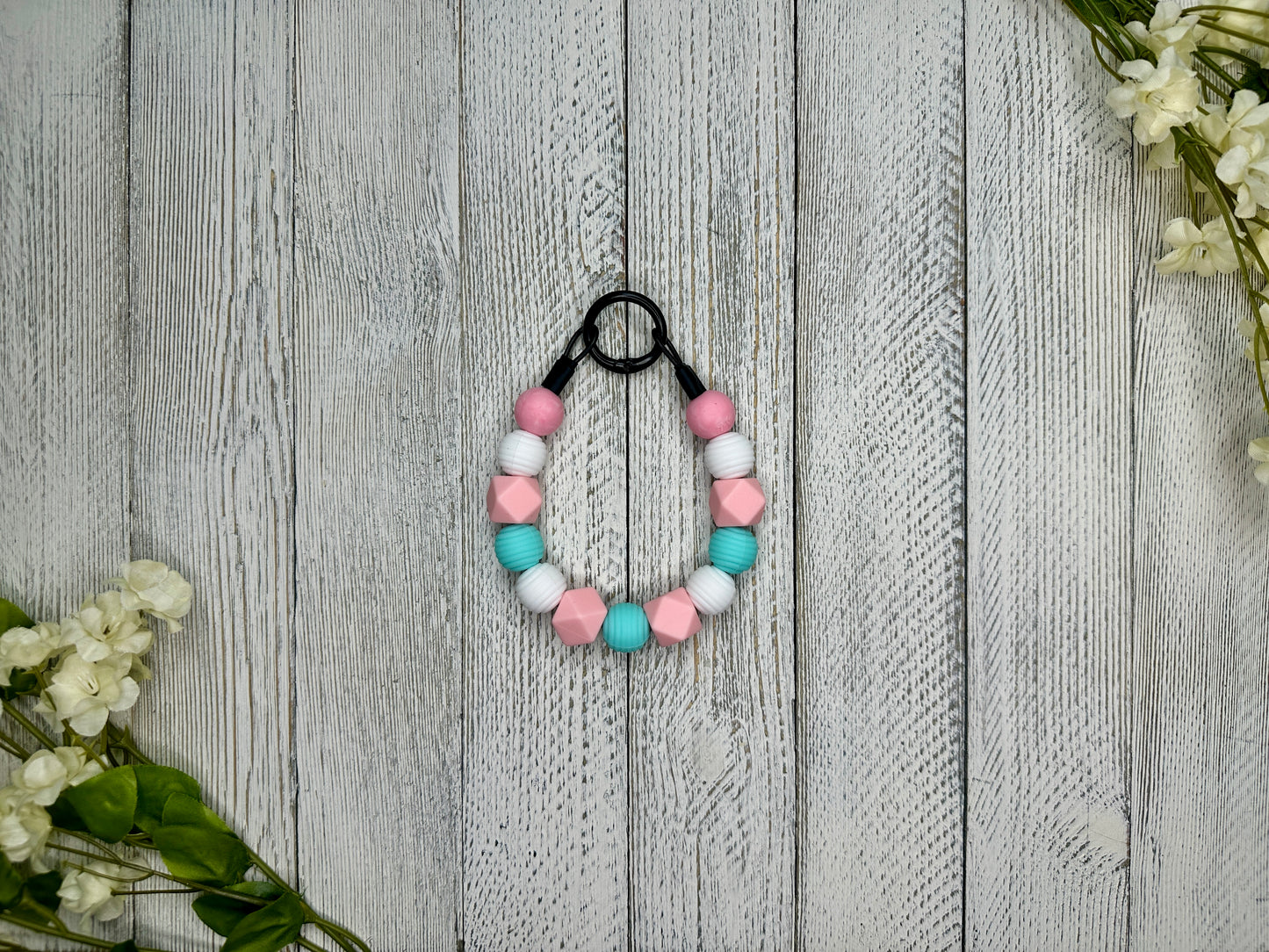 Beaded dog necklace with a pink, blue, and white pattern. Displayed on a wooden surface with flowers for decoration. 