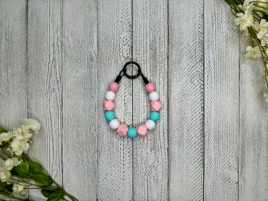 Beaded dog necklace with a pink, blue, and white pattern. Displayed on a wooden surface with flowers for decoration. 