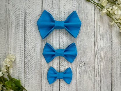 Three pet bow ties (small, medium, large) that are bright blue, displayed on a wooden surface with flowers for decoration.