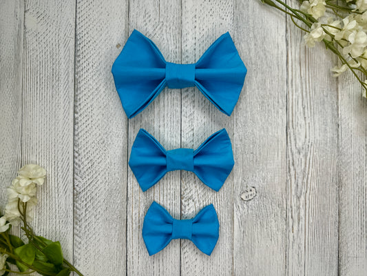 Three pet bow ties (small, medium, large) that are bright blue, displayed on a wooden surface with flowers for decoration.
