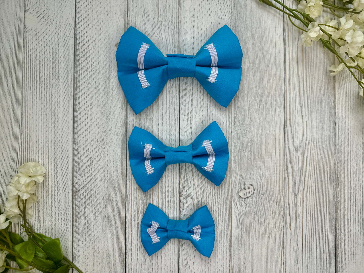 Picture of the backside of pet bow ties showing the elastic straps that fit around a collar. Displayed on a wooden surface with flowers for decoration. 