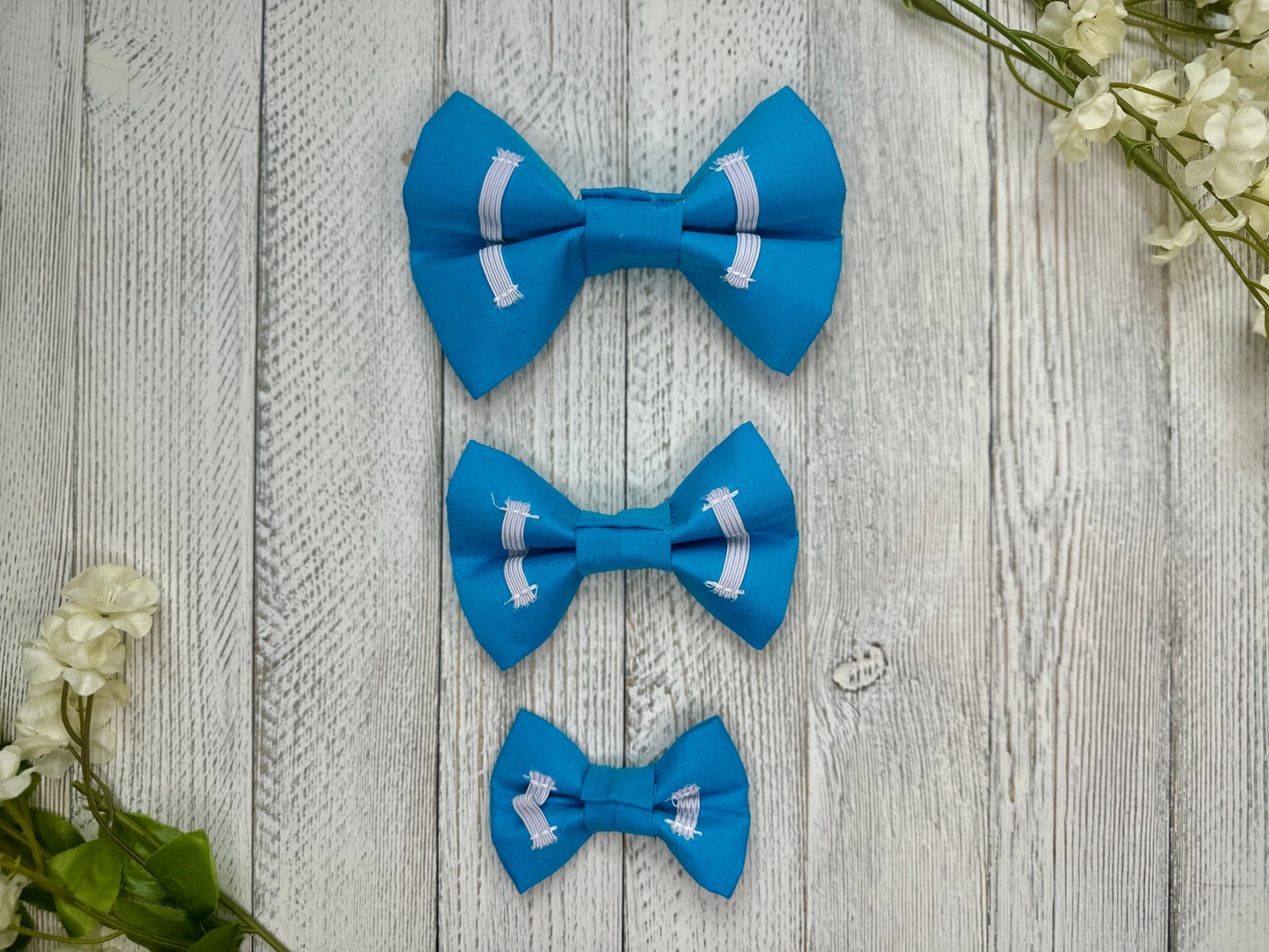 Picture of the backside of pet bow ties showing the elastic straps that fit around a collar. Displayed on a wooden surface with flowers for decoration. 