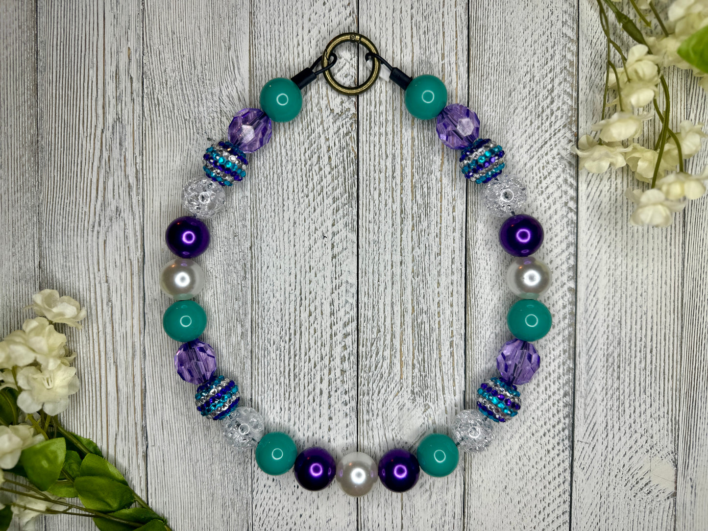 Beaded pet necklace with a purple and teal pattern, including sparkles. Displayed on a wooden surface with flowers for decoration. 