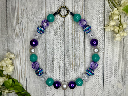 Beaded pet necklace with a purple and teal pattern, including sparkles. Displayed on a wooden surface with flowers for decoration. 