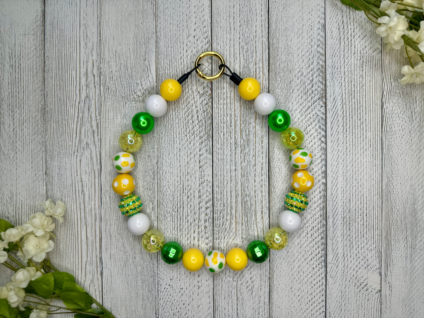 Beaded pet necklace with a green, yellow, and white pattern, including lemons and limes and polka dots. Displayed on a wooden surface with flowers for decoration. 