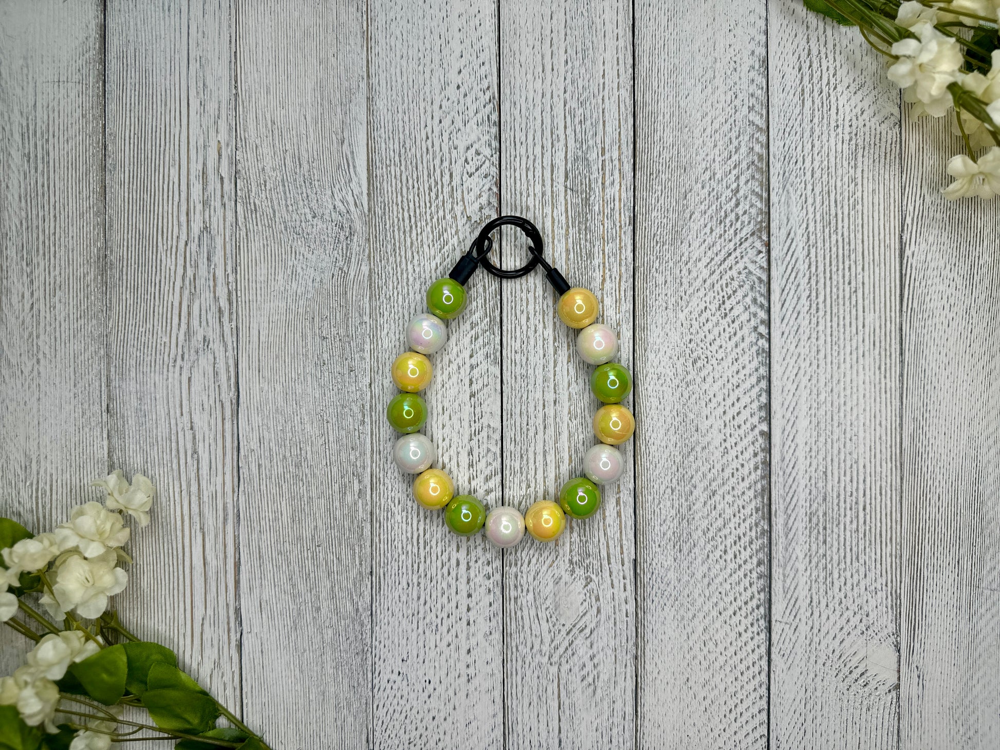 Beaded pet necklace with a green, yellow, and white pattern. Displayed on a wooden surface with flowers for decoration. 