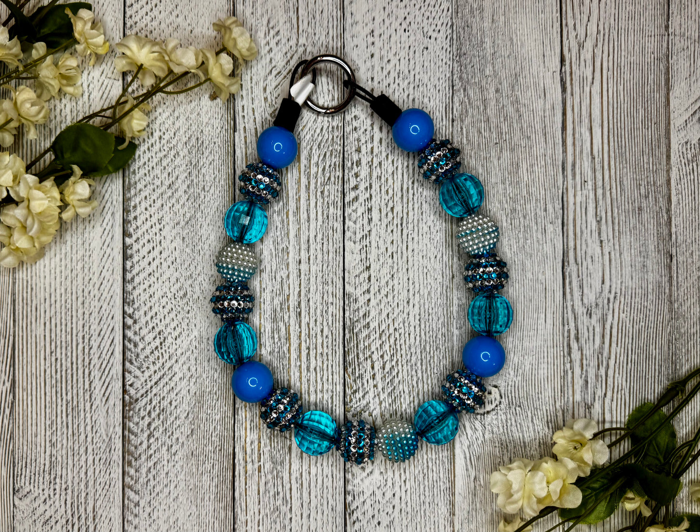 Beaded dog necklace with a light blue and silver pattern including stripes. Displayed on a wooden surface with flowers for decoration.
