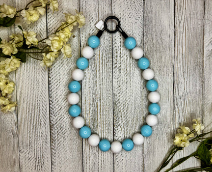 Beaded dog necklace with a light blue and white pattern. Displayed on a wooden surface with flowers for decoration.