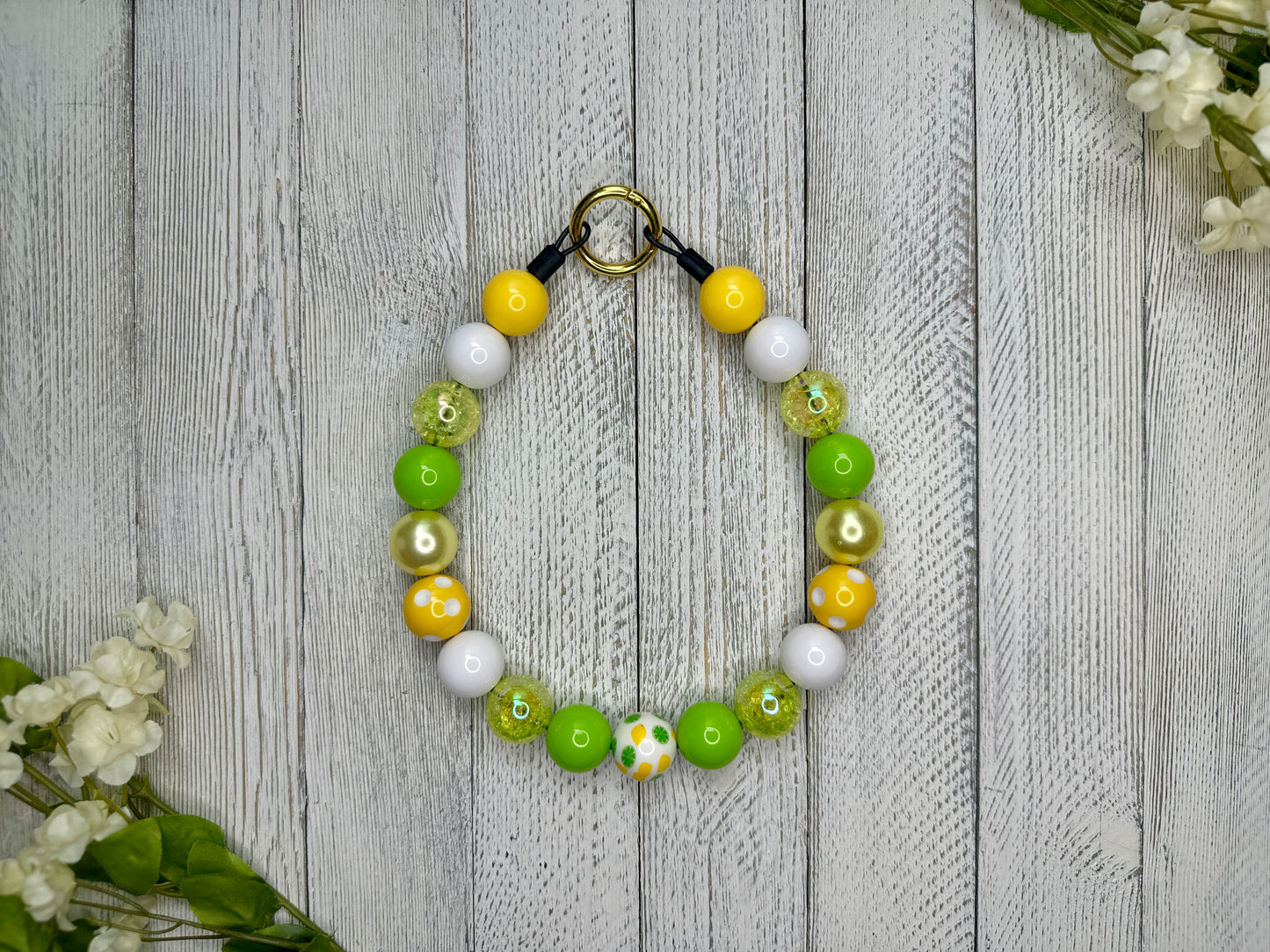 Beaded pet necklace with a green, yellow, and white pattern, including lemons and limes and polka dots. Displayed on a wooden surface with flowers for decoration. 