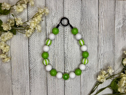 Beaded dog necklace with a lime green and white pattern including stripes. Displayed on a wooden surface with flowers for decoration.