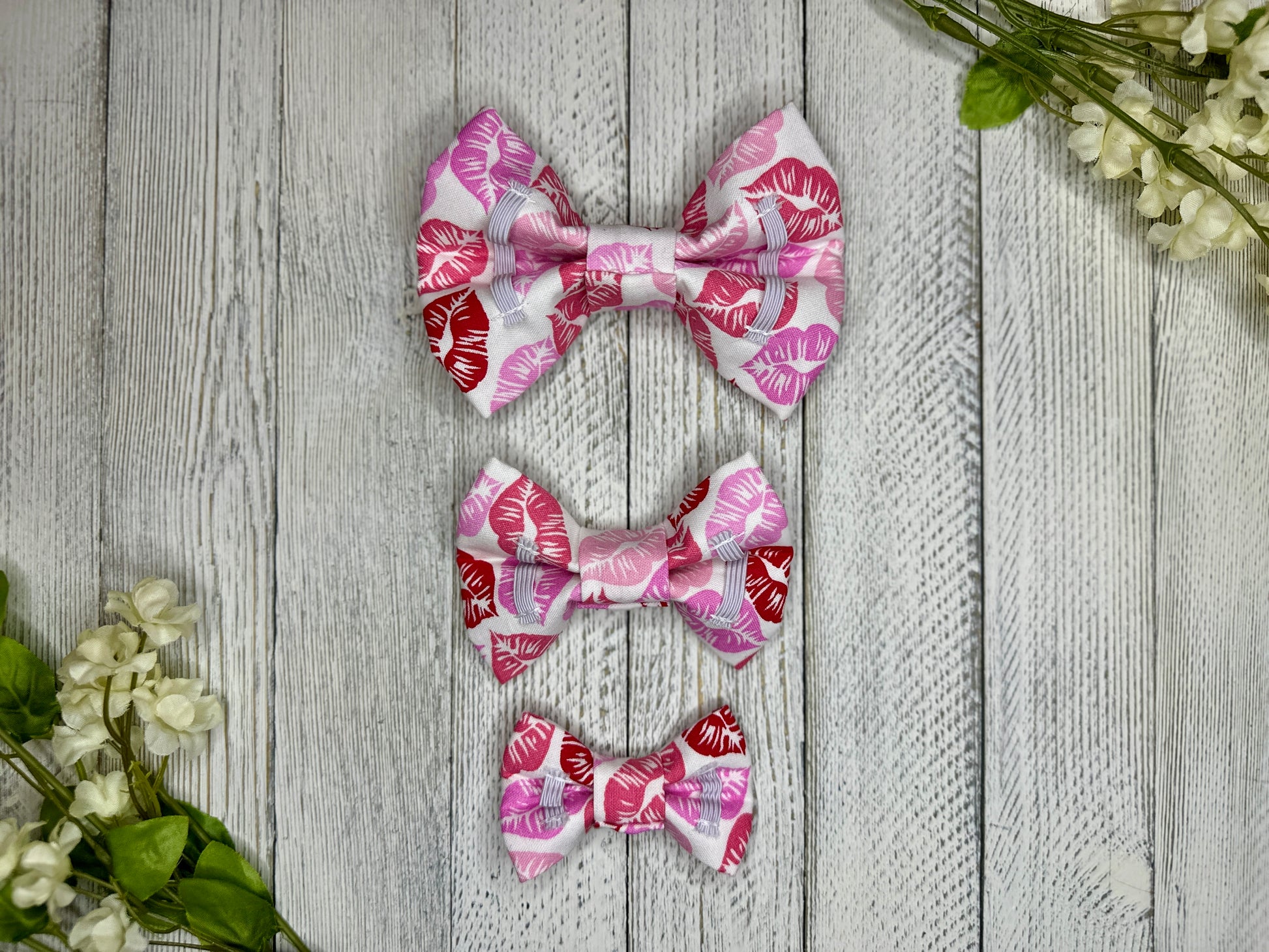 Picture of the backside of pet bow ties showing the elastic straps that fit around a collar. Displayed on a wooden surface with flowers for decoration. 