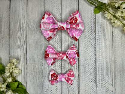 Three pet bow ties (small, medium, large) with red and pink lipstick kisses displayed on a wooden surface with flowers for decoration. 