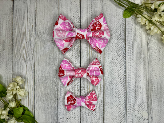 Three pet bow ties (small, medium, large) with red and pink lipstick kisses displayed on a wooden surface with flowers for decoration. 