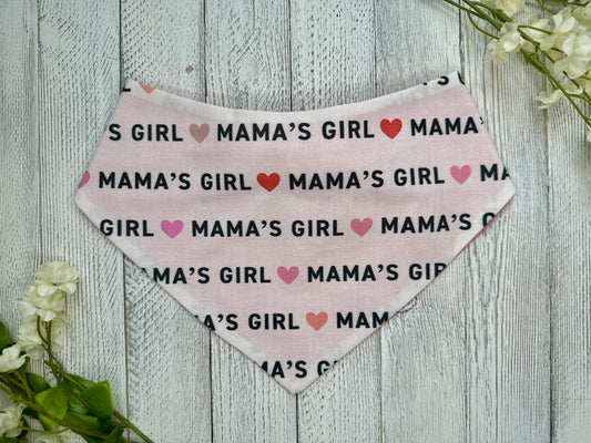 A pet bandana with a white background displaying "Mama's Girl" with hearts, displayed on a wooden surface with flowers for decoration. 