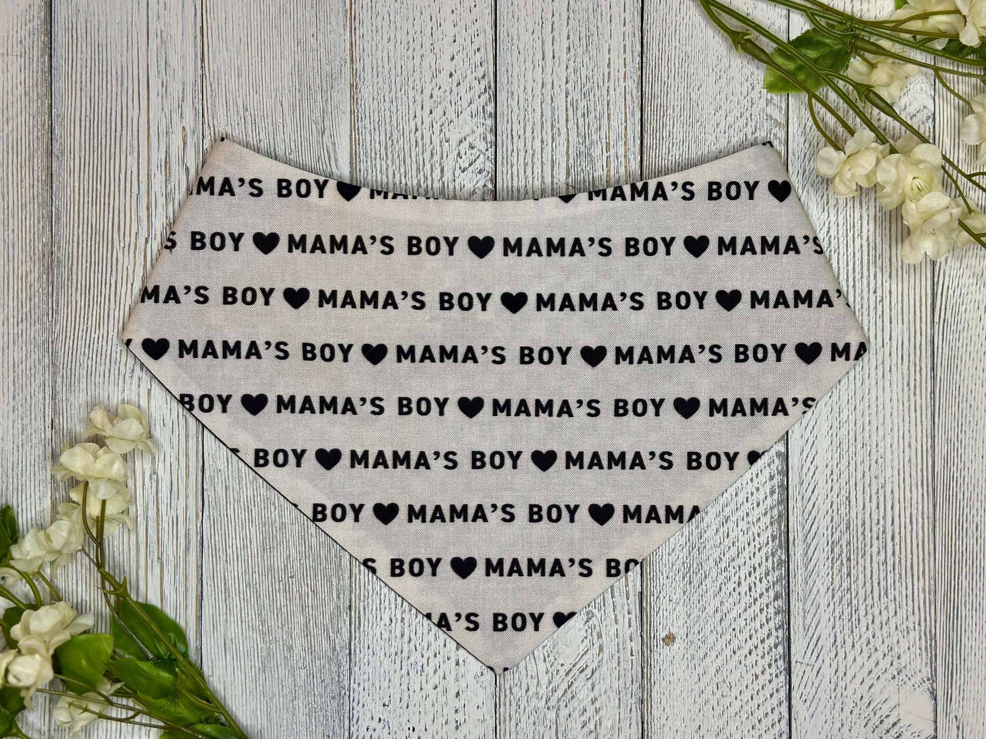 A pet bandana with a off white background featuring a mama's boy pattern with hearts, displayed on a wooden surface with flowers for decoration. 