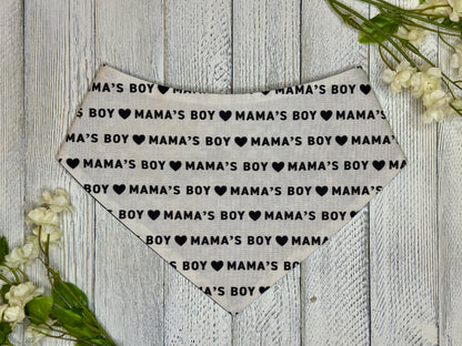 A pet bandana with a off white background featuring a mama's boy pattern with hearts, displayed on a wooden surface with flowers for decoration. 