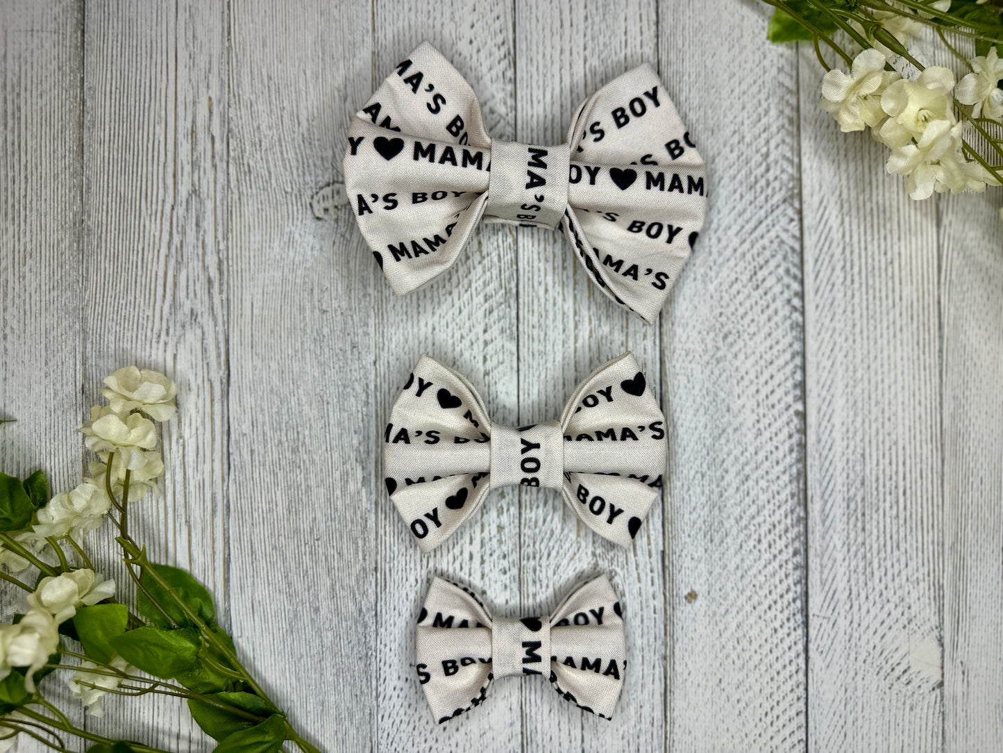 Three pet bow ties (small, medium, large) with an off white background, mama's boy pattern, and hearts, displayed on a wooden surface with flowers for decoration. 