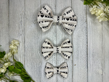 Three pet bow ties (small, medium, large) with an off white background, mama's boy pattern, and hearts, displayed on a wooden surface with flowers for decoration. 