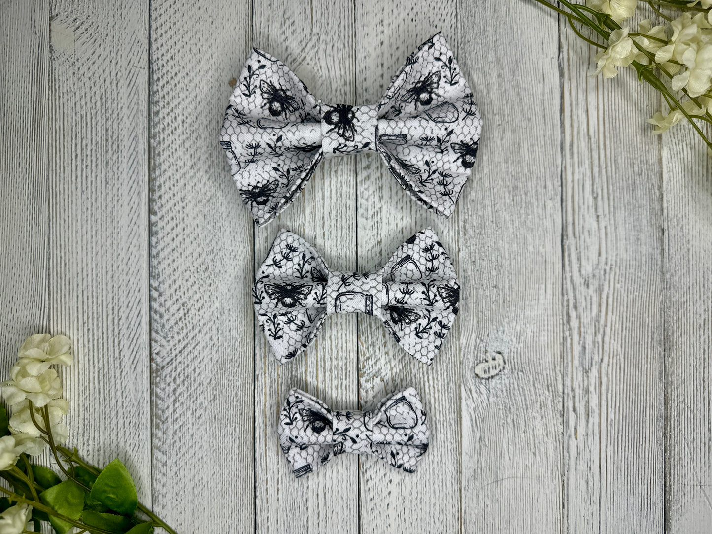Three pet bow ties (small, medium, large) with a white background, mason jars, and bees, displayed on a wooden surface with flowers for decoration. 