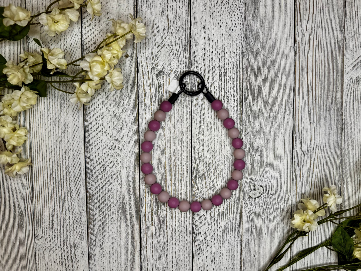 Beaded dog necklace with a light and dark mauve pattern. Displayed on a wooden surface with flowers for decoration.