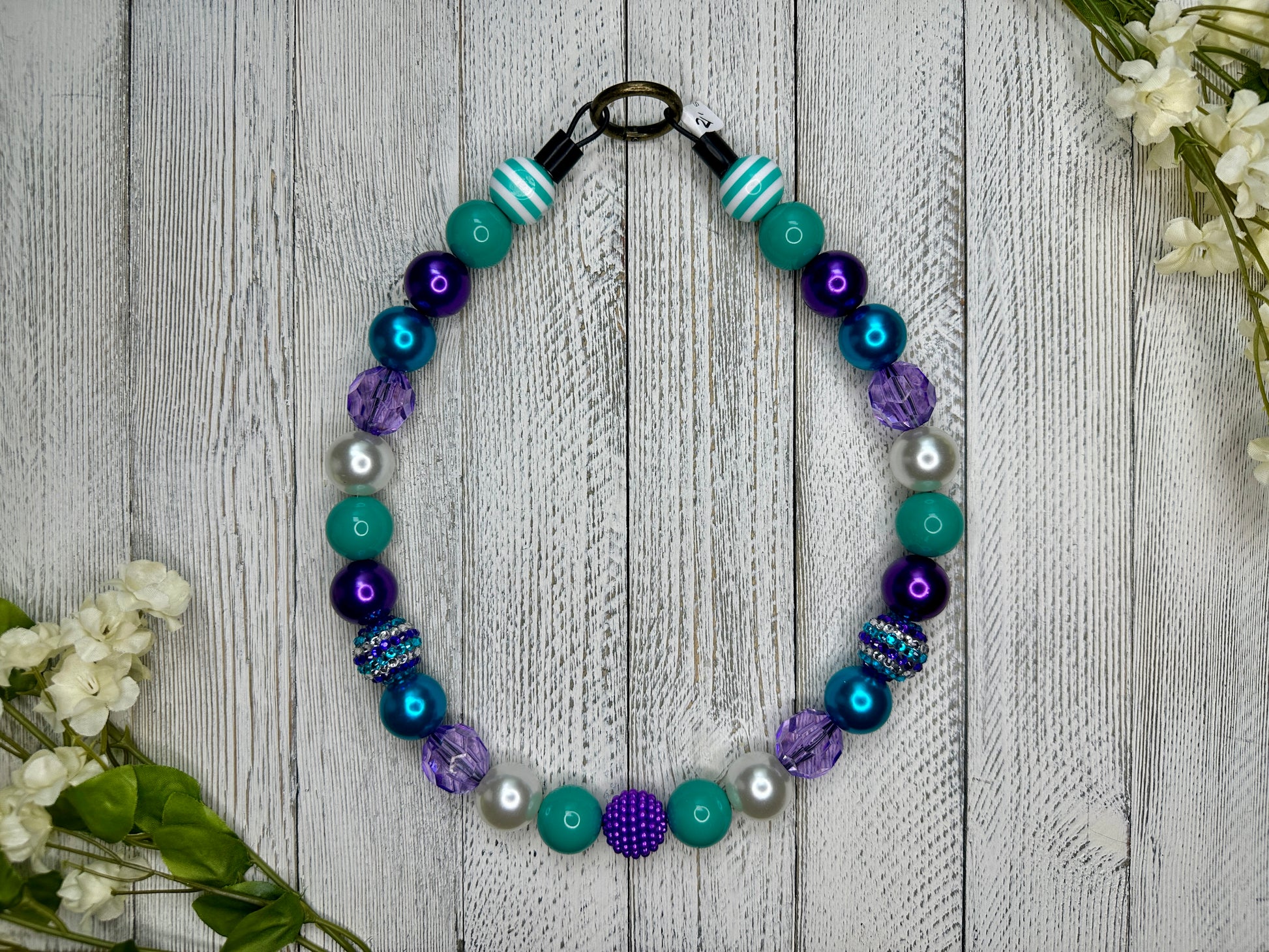 Beaded pet necklace with a purple and teal pattern, including stripes. Displayed on a wooden surface with flowers for decoration. 