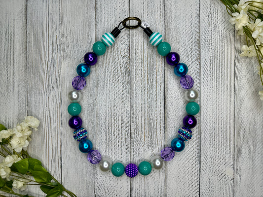 Beaded pet necklace with a purple and teal pattern, including stripes. Displayed on a wooden surface with flowers for decoration. 