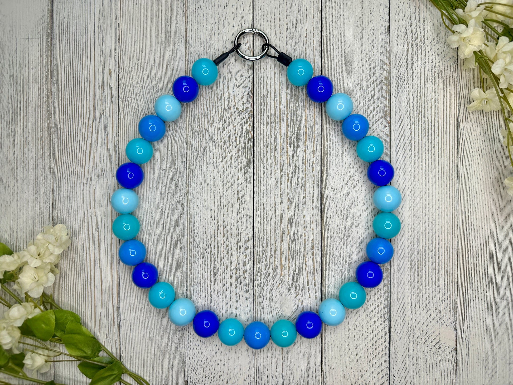 Beaded pet necklace with a a variety of blues pattern. Displayed on a wooden surface with flowers for decoration. 