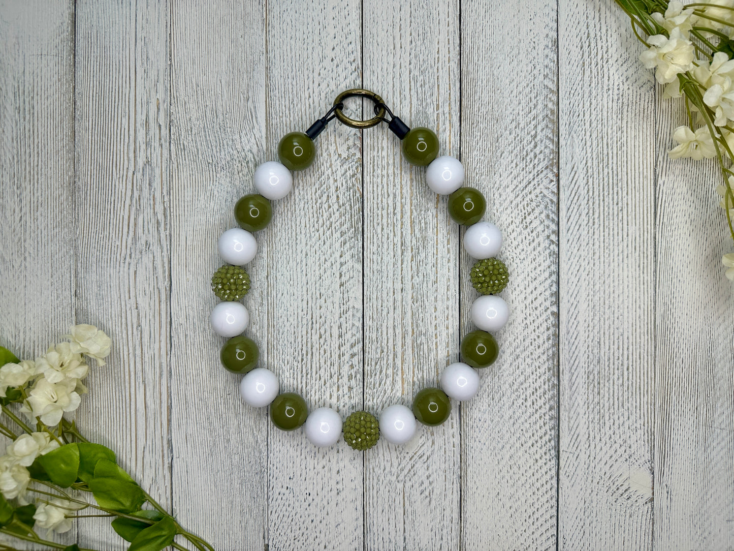 Beaded pet necklace with an olive and white pattern, including sparkles. Displayed on a wooden surface with flowers for decoration. 