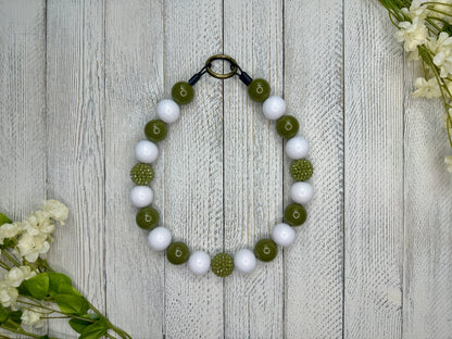 Beaded pet necklace with an olive and white pattern, including sparkles. Displayed on a wooden surface with flowers for decoration. 