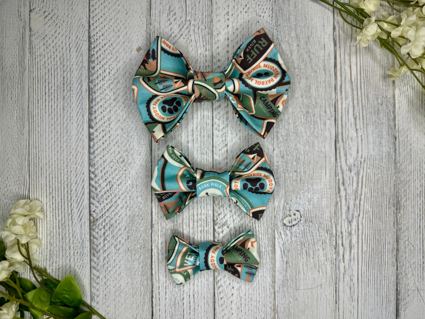 Three pet bow ties (small, medium, large) with a blue background and pup ranger patch pattern displayed on a wooden surface with flowers for decoration. 