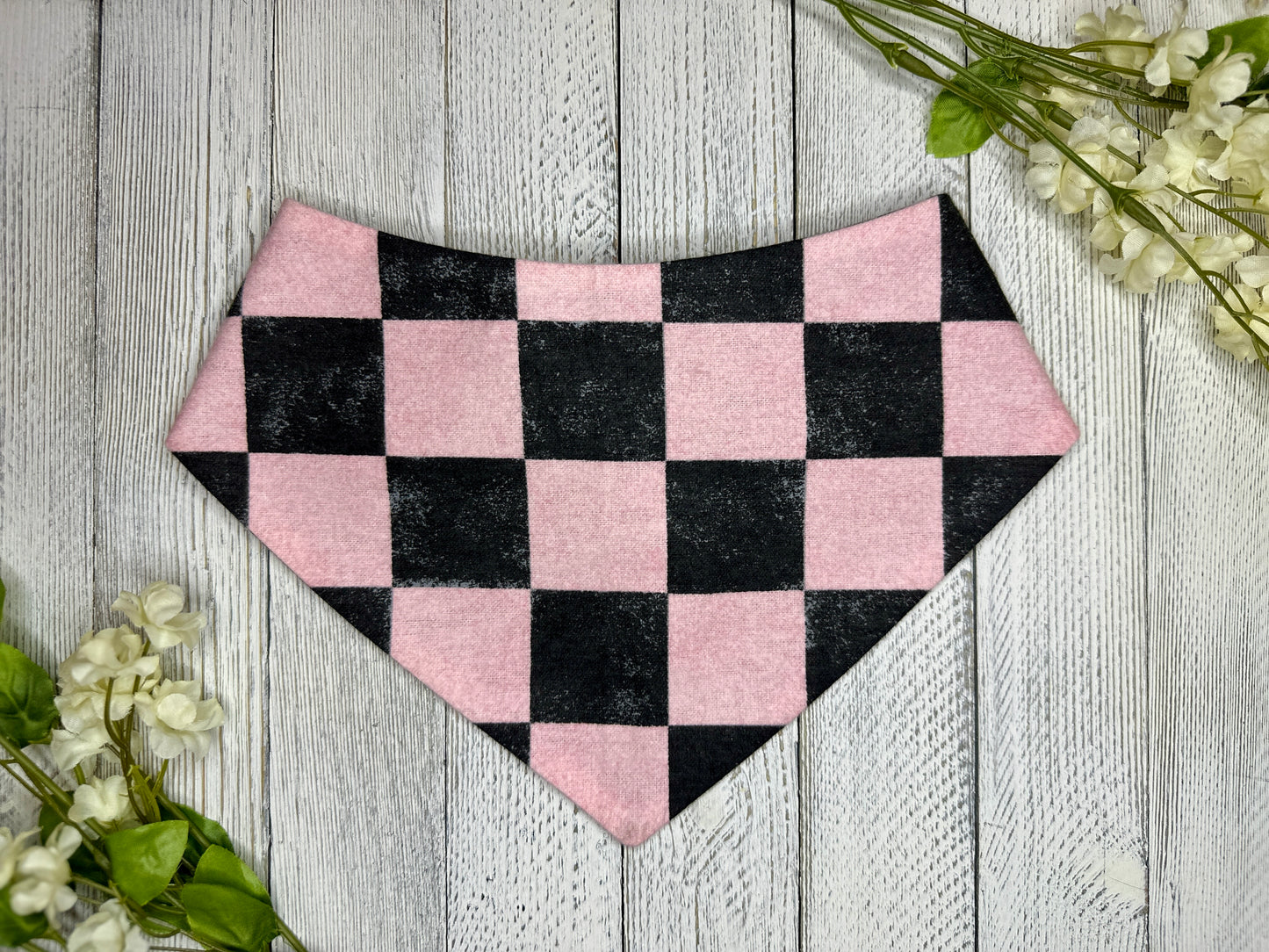 A pet bandana with a black and pink checkered pattern, displayed on a wooden surface with flowers for decoration. 