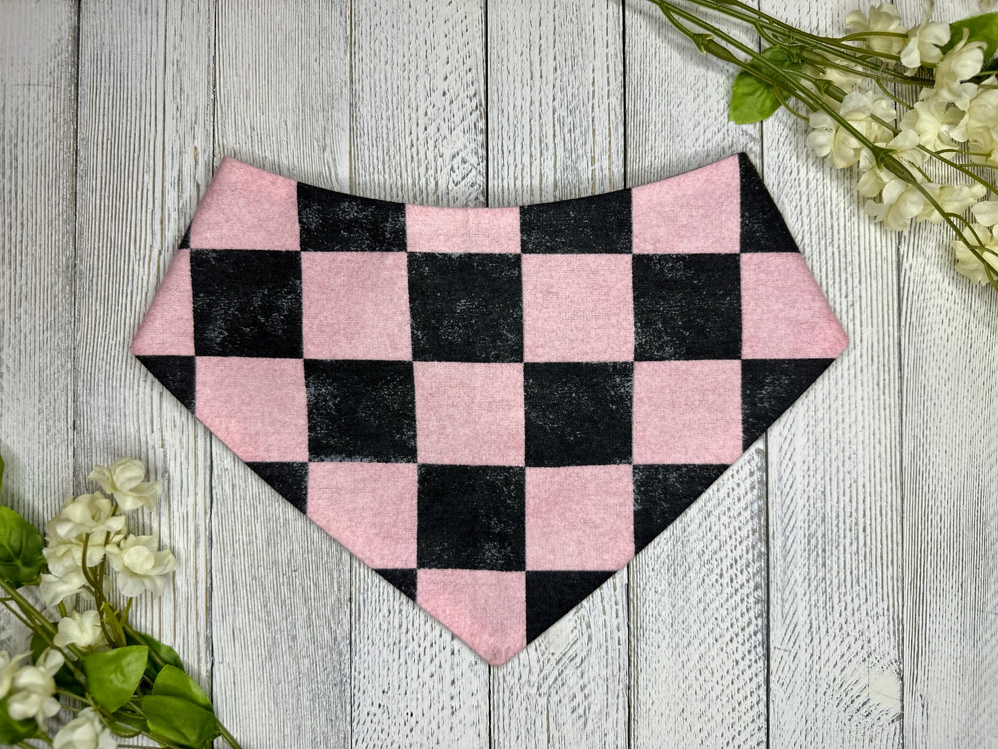 A pet bandana with a black and pink checkered pattern, displayed on a wooden surface with flowers for decoration. 