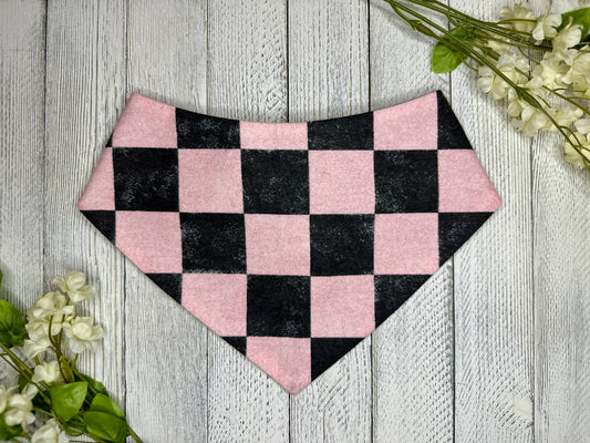 A pet bandana with a black and pink checkered pattern, displayed on a wooden surface with flowers for decoration. 