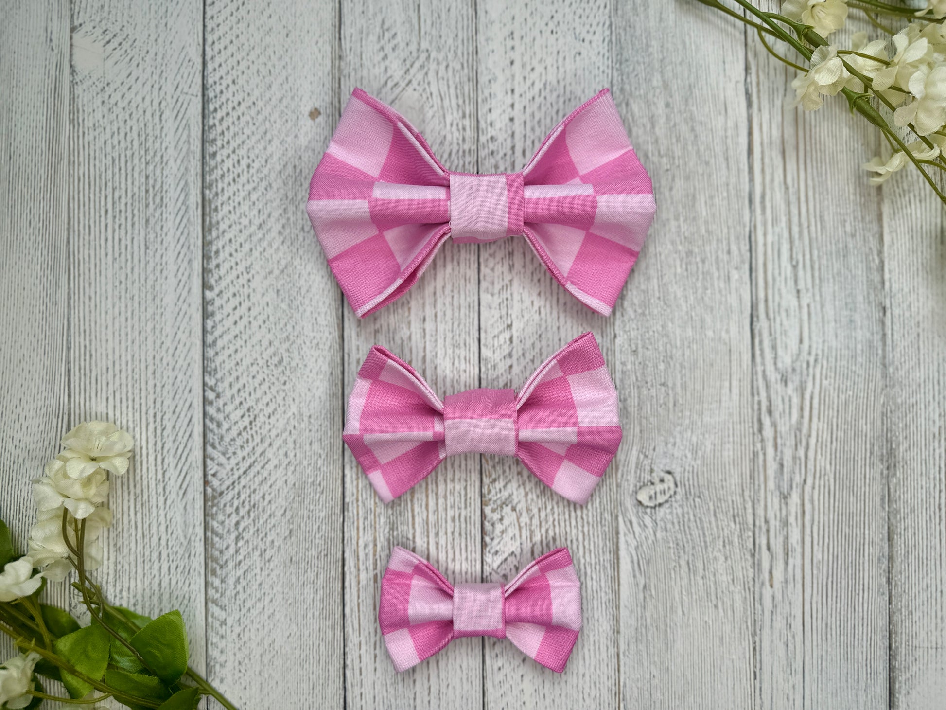 Three pet bow ties (small, medium, large) with an pink checkered pattern, displayed on a wooden surface with flowers for decoration. 