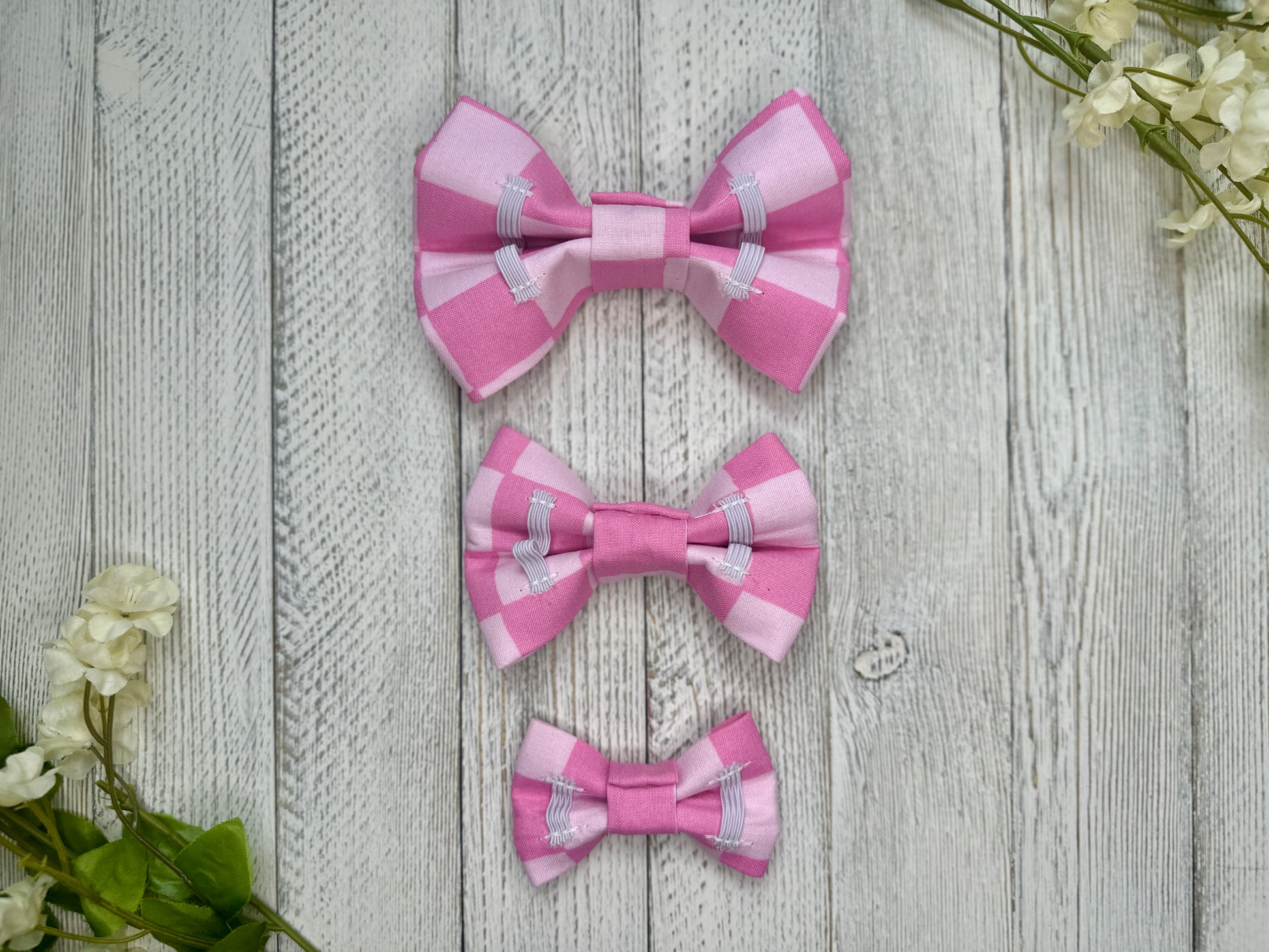 Picture of the backside of pet bow ties showing the elastic straps that fit around a collar. Displayed on a wooden surface with flowers for decoration. 
