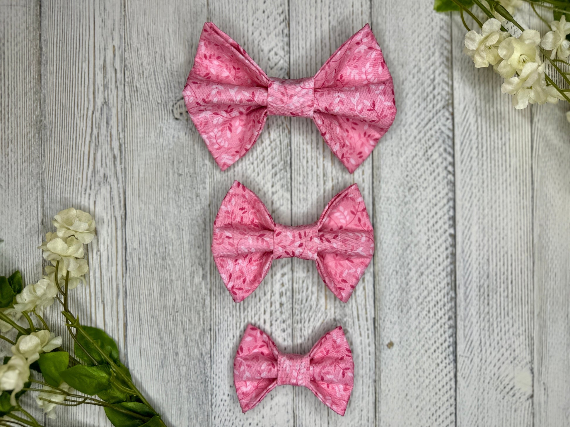 Three pet bow ties (small, medium, large) with a pink background and subtle darker and lighter pink vines, displayed on a wooden surface with flowers for decoration.