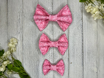 Three pet bow ties (small, medium, large) with a pink background and subtle darker and lighter pink vines, displayed on a wooden surface with flowers for decoration.
