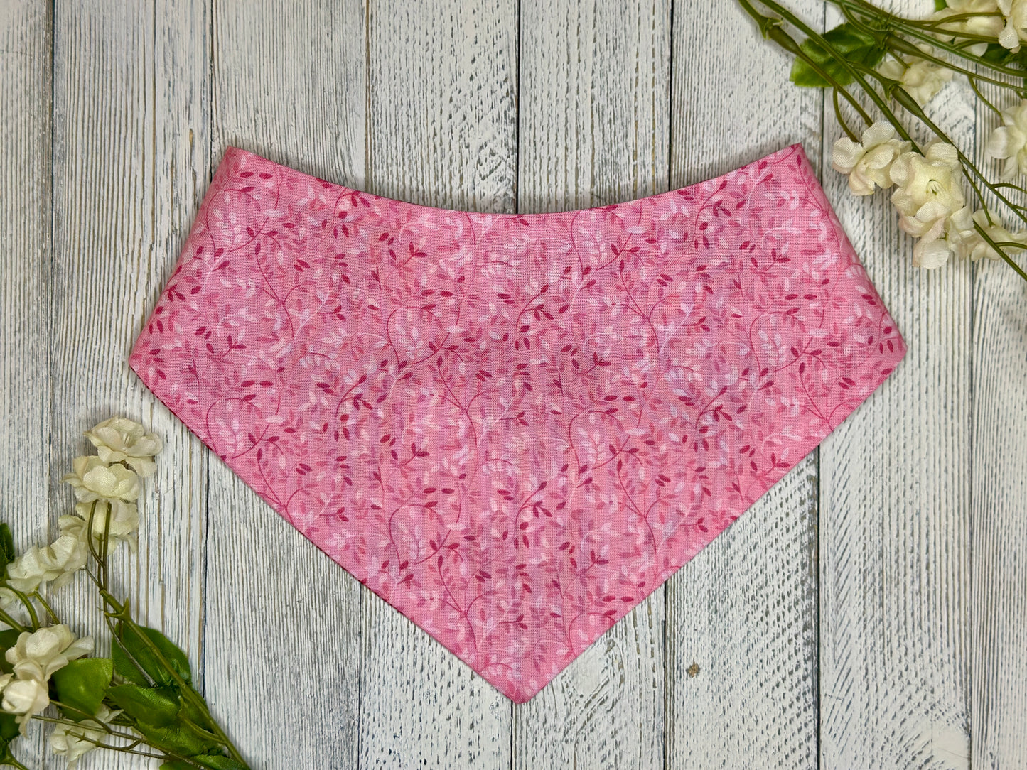 A pet bandana with a pink background featuring a subtle pi, displayed on a wooden surface with flowers for decoration. k vine pattern