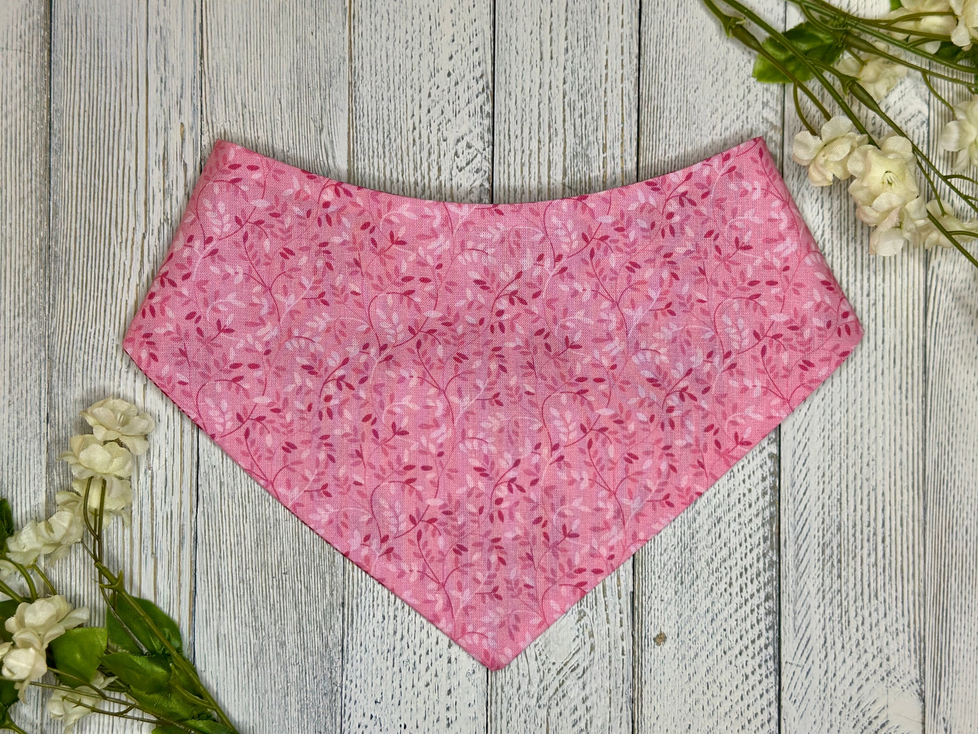 A pet bandana with a pink background featuring a subtle pi, displayed on a wooden surface with flowers for decoration. k vine pattern