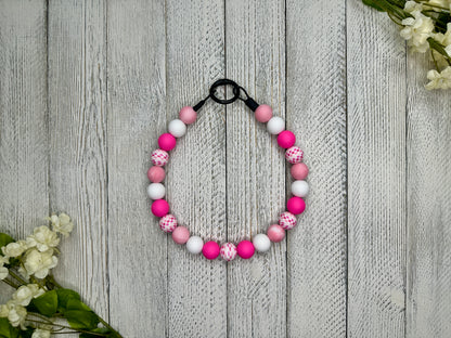 Beaded pet necklace with a pink and white pattern including checkers. Displayed on a wooden surface with flowers for decoration. 