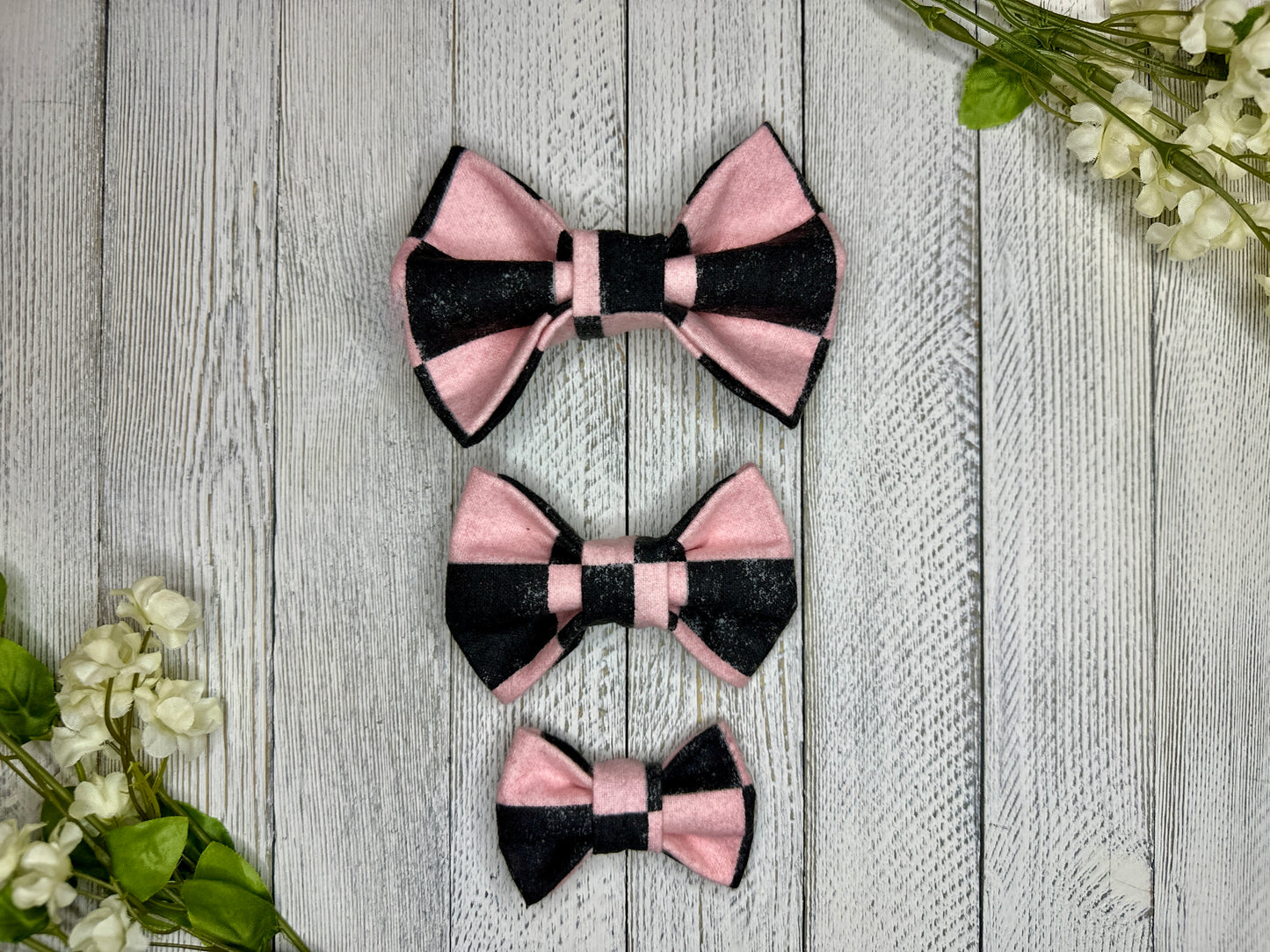 Three pet bow ties (small, medium, large) with a pink and black checkered pattern displayed on a wooden surface with flowers for decoration.