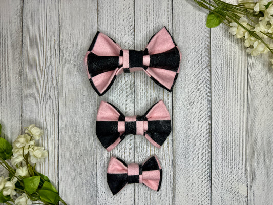 Three pet bow ties (small, medium, large) with a pink and black checkered pattern displayed on a wooden surface with flowers for decoration.