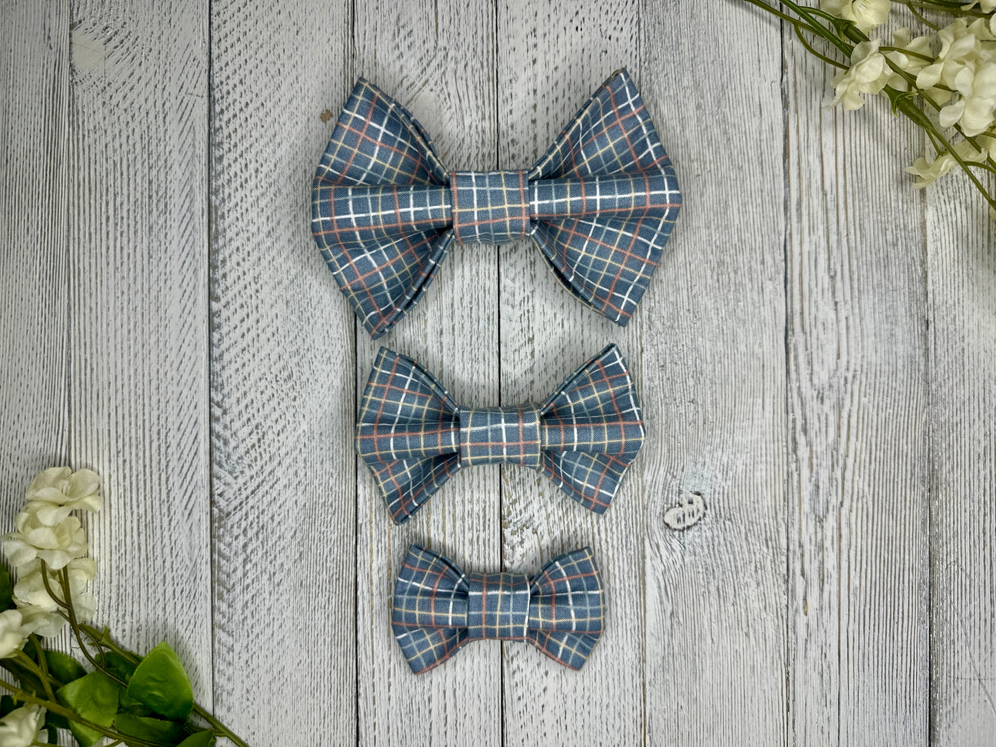 Three pet bow ties (small, medium, large) with a plaid pattern displayed on a wooden surface with flowers for decoration. 