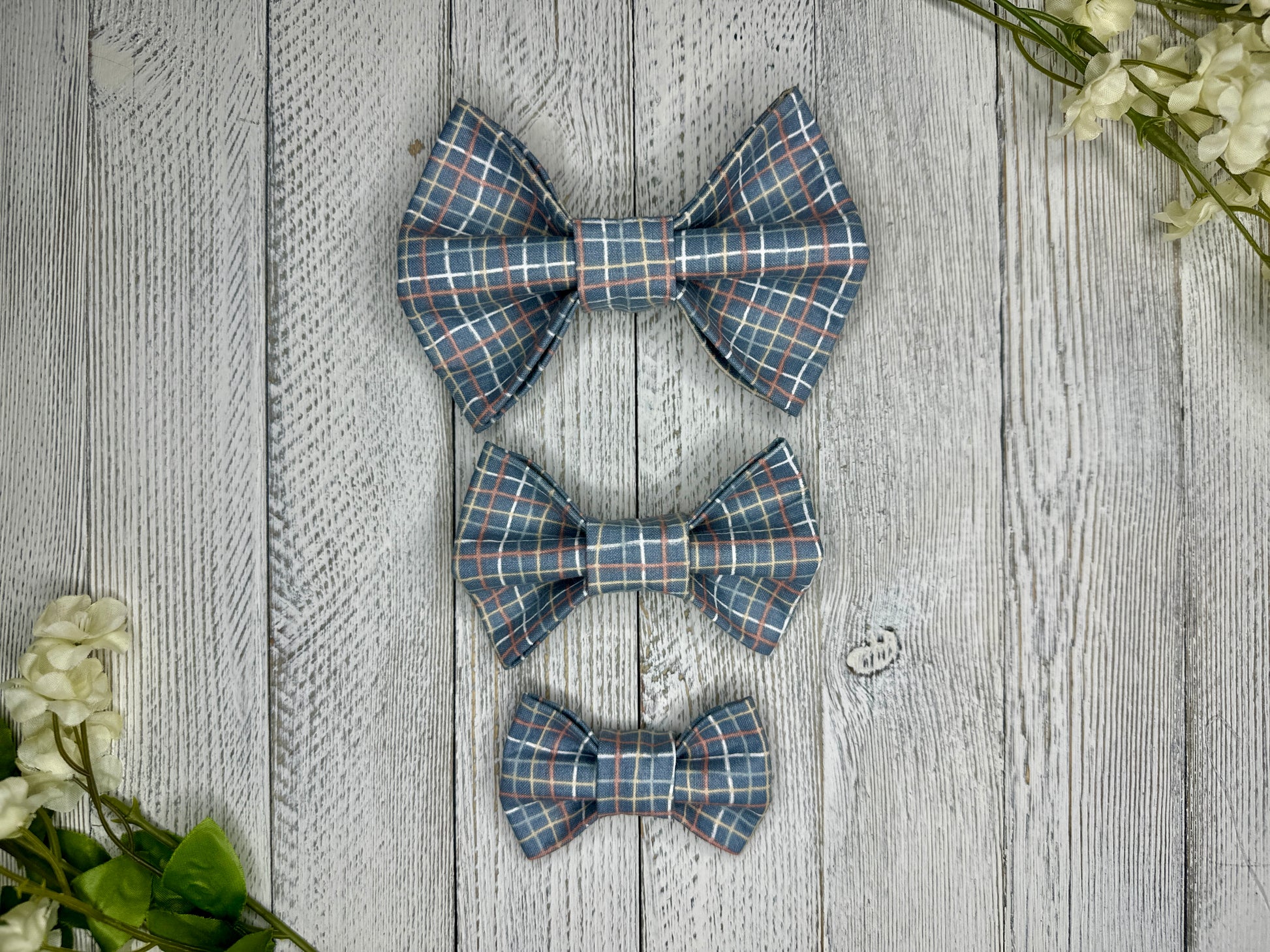 Three pet bow ties (small, medium, large) with a plaid pattern displayed on a wooden surface with flowers for decoration. 
