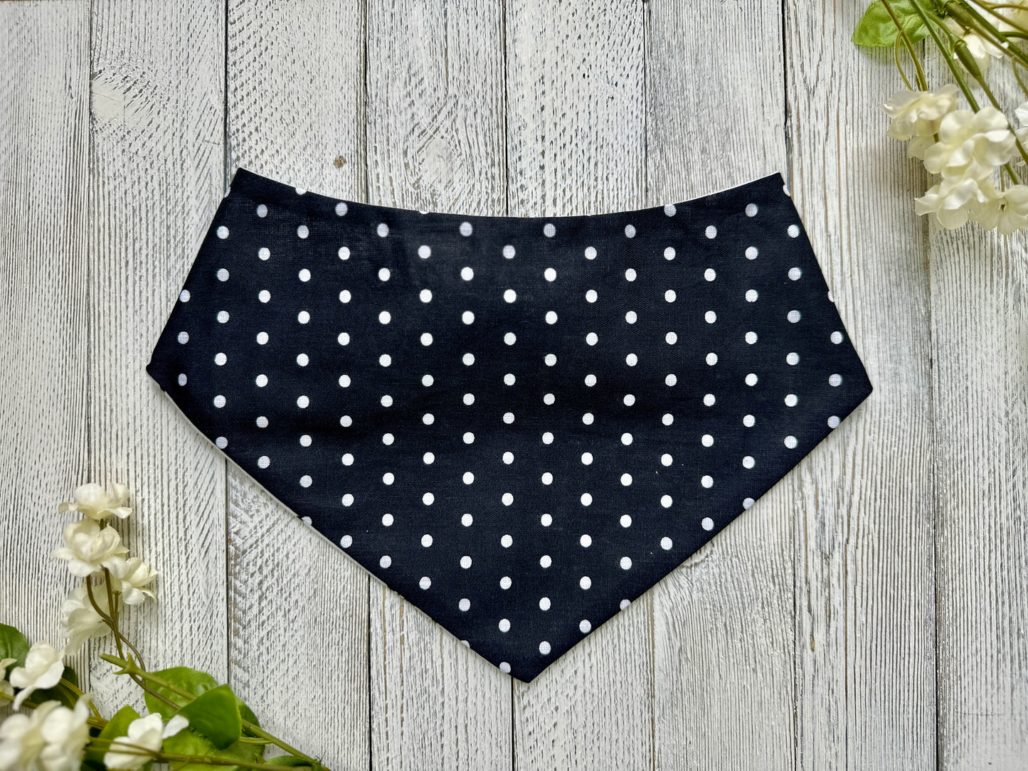 A pet bandana with a black background featuring a white polka dot pattern, displayed on a wooden surface with flowers for decoration. 