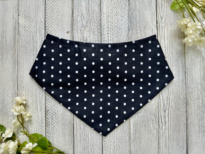 A pet bandana with a black background featuring a white polka dot pattern, displayed on a wooden surface with flowers for decoration. 
