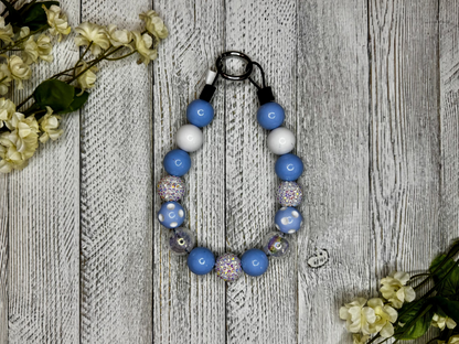 Beaded dog necklace with a light blue and white pattern including polka dots. Displayed on a wooden surface with flowers for decoration.