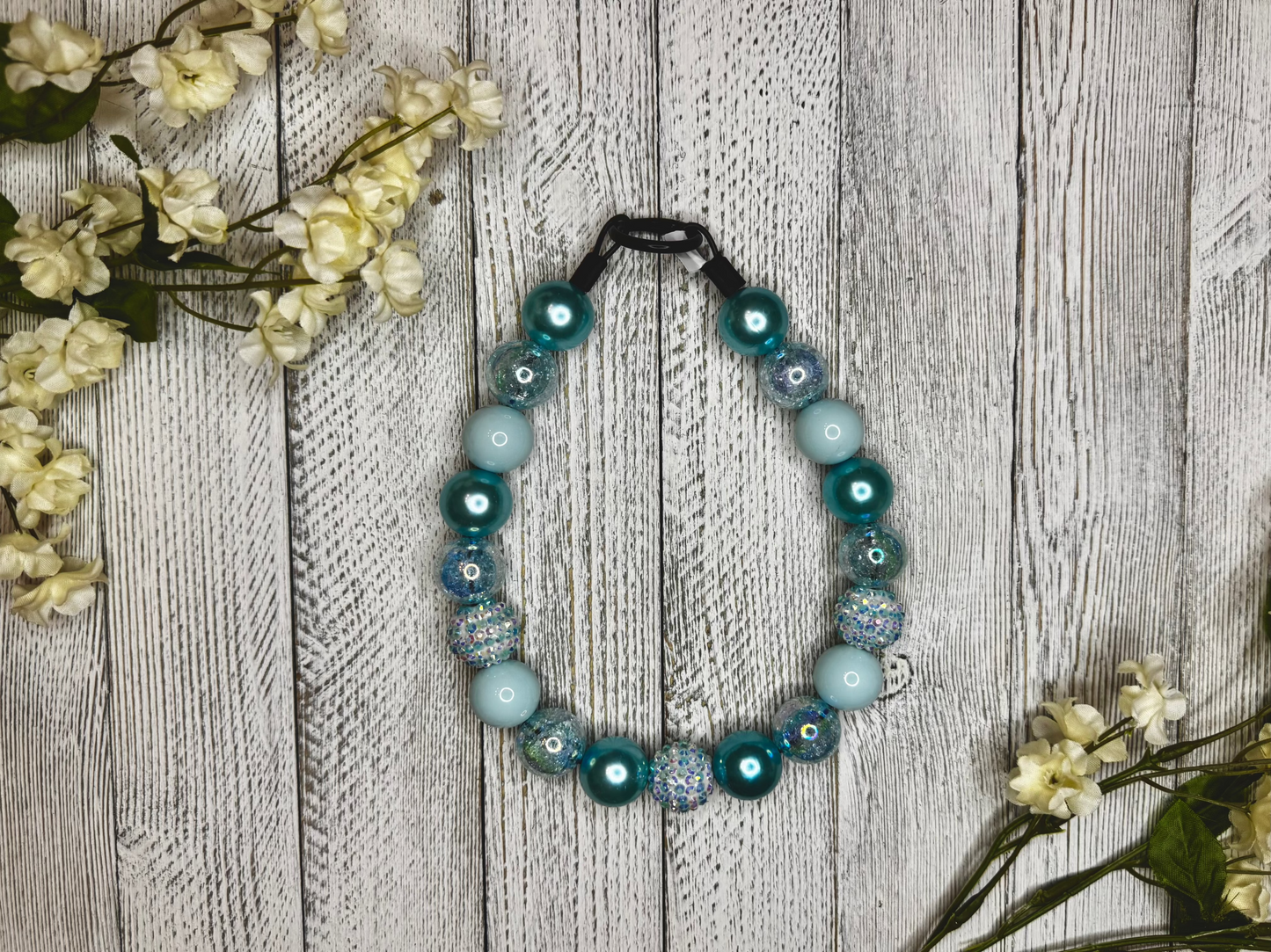 Beaded dog necklace with a light blue pattern including stripes. Displayed on a wooden surface with flowers for decoration.
