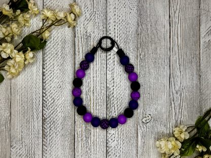 Beaded dog necklace with a purple and black pattern including checkers. Displayed on a wooden surface with flowers for decoration.